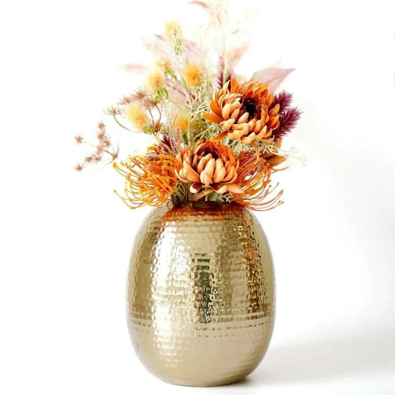 Gold hammered oval vase with an orange floral arrangement on a white background