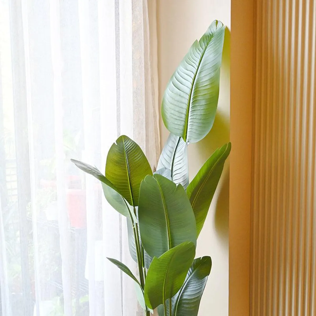 Tall artificial leafy plant placed beside a bright window and wooden wall