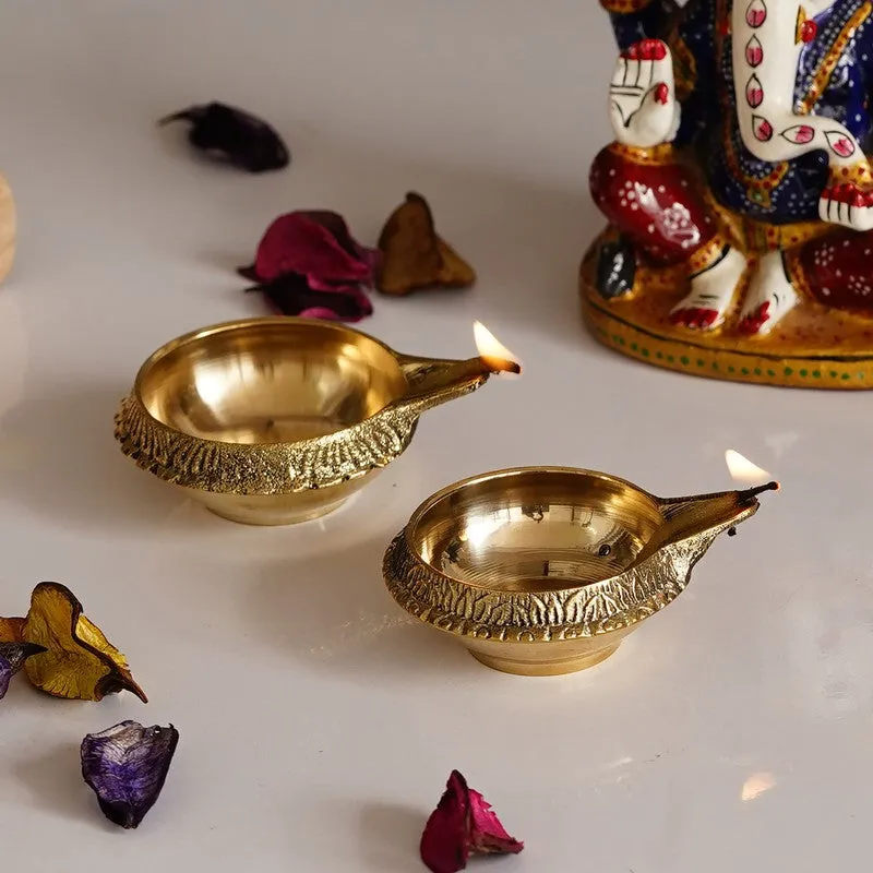 Two lit gold brass diyas with textured rims on a white surface
