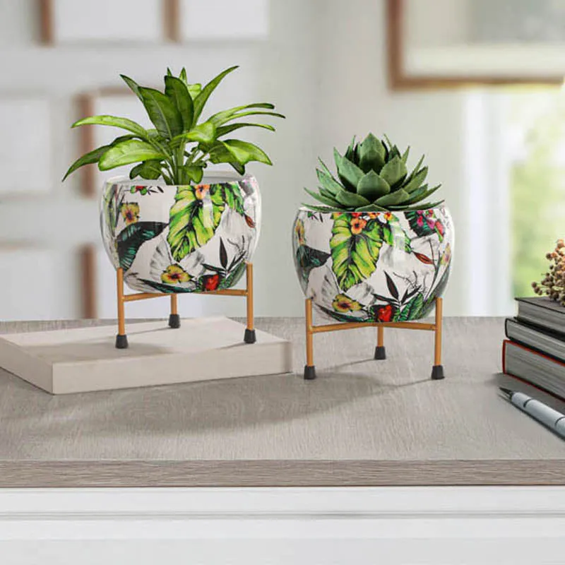Two tropical print planters on stands styled with green plants on a tabletop