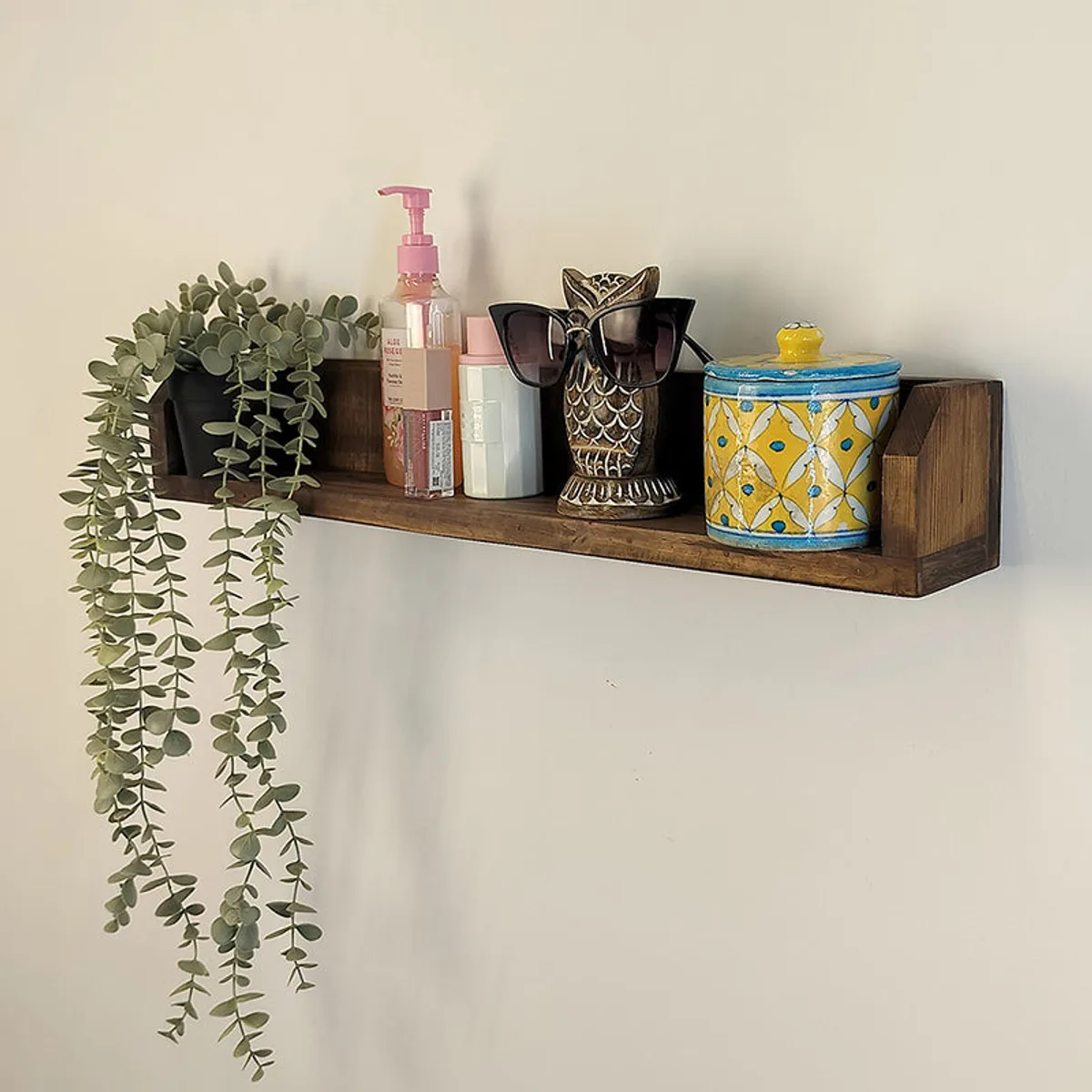 Angled view of a brown wooden wall shelf styled with a plant, toiletries, figurine and lidded box.