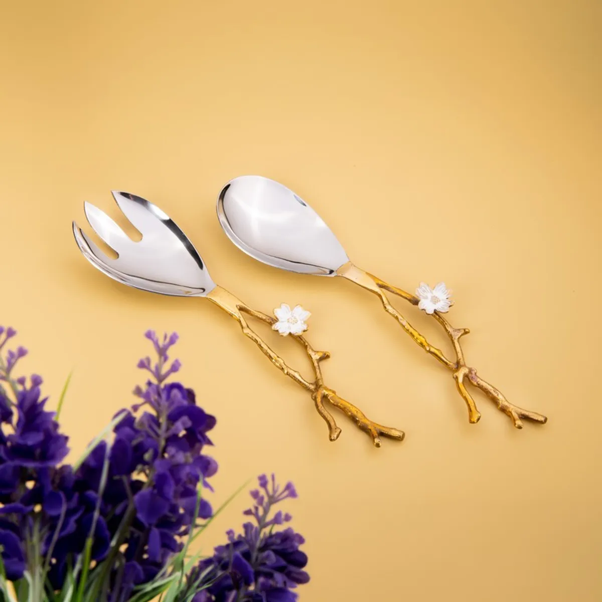 Two silver and gold salad servers with white floral details, styled beside purple flowers.
