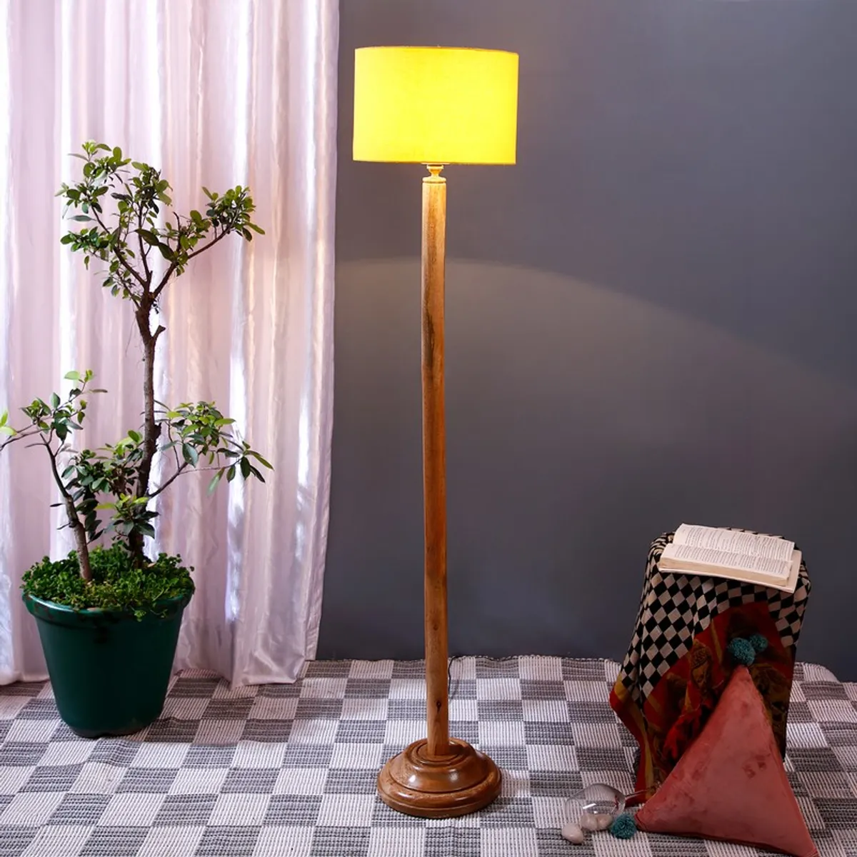Wooden floor lamp with yellow drum shade placed beside a potted plant and side stool