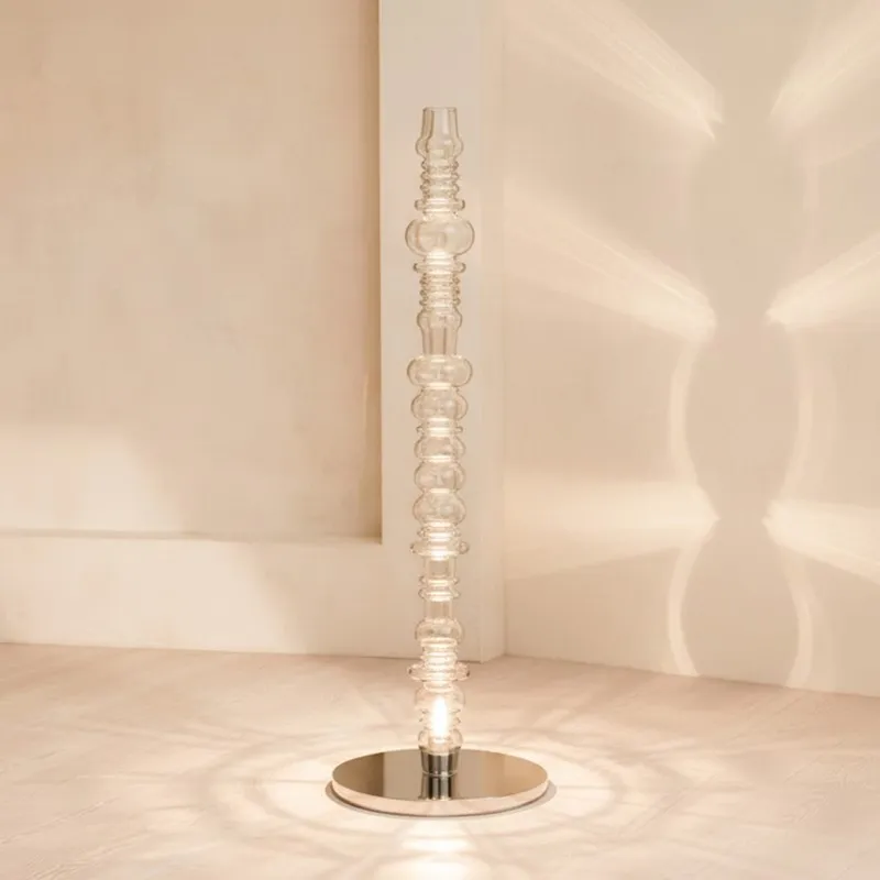 Tall clear glass floor lamp casting soft light against a wall