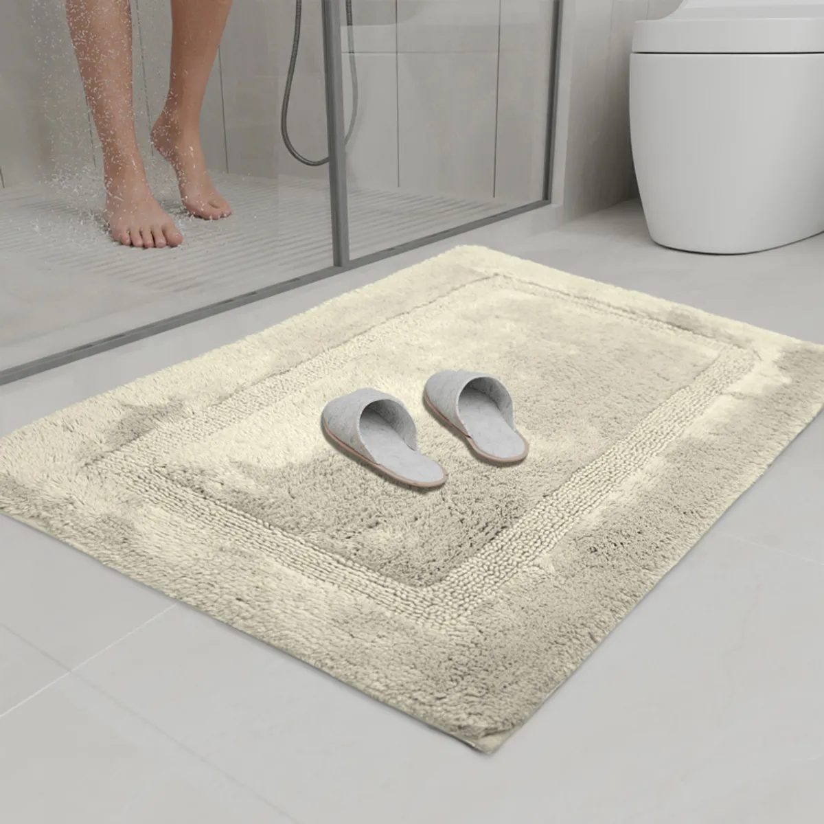 Grey bath mat positioned outside a glass shower enclosure with slippers on top.
