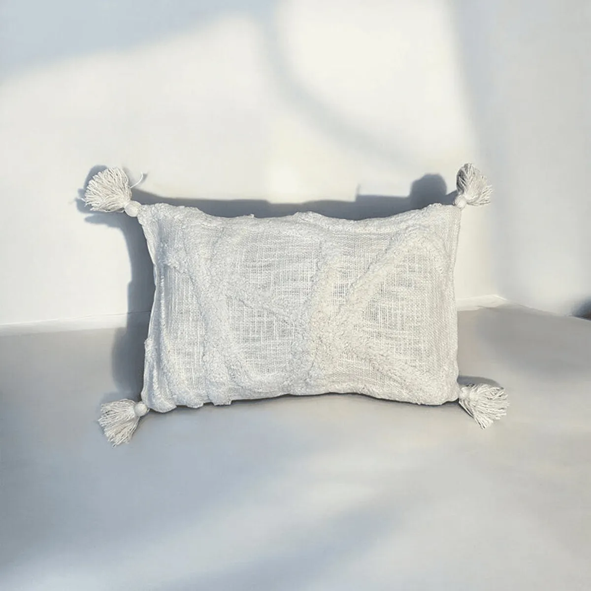 White rectangular tufted cushion cover with tassels against a softly lit plain backdrop