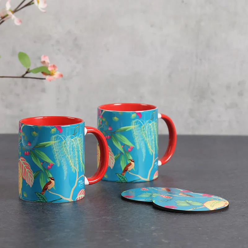 Two blue ceramic mugs with red handles and tropical bird-and-leaf designs beside matching coasters