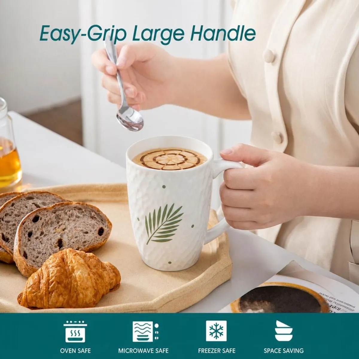 Hand holding a white leaf-print mug beside bread and a spoon