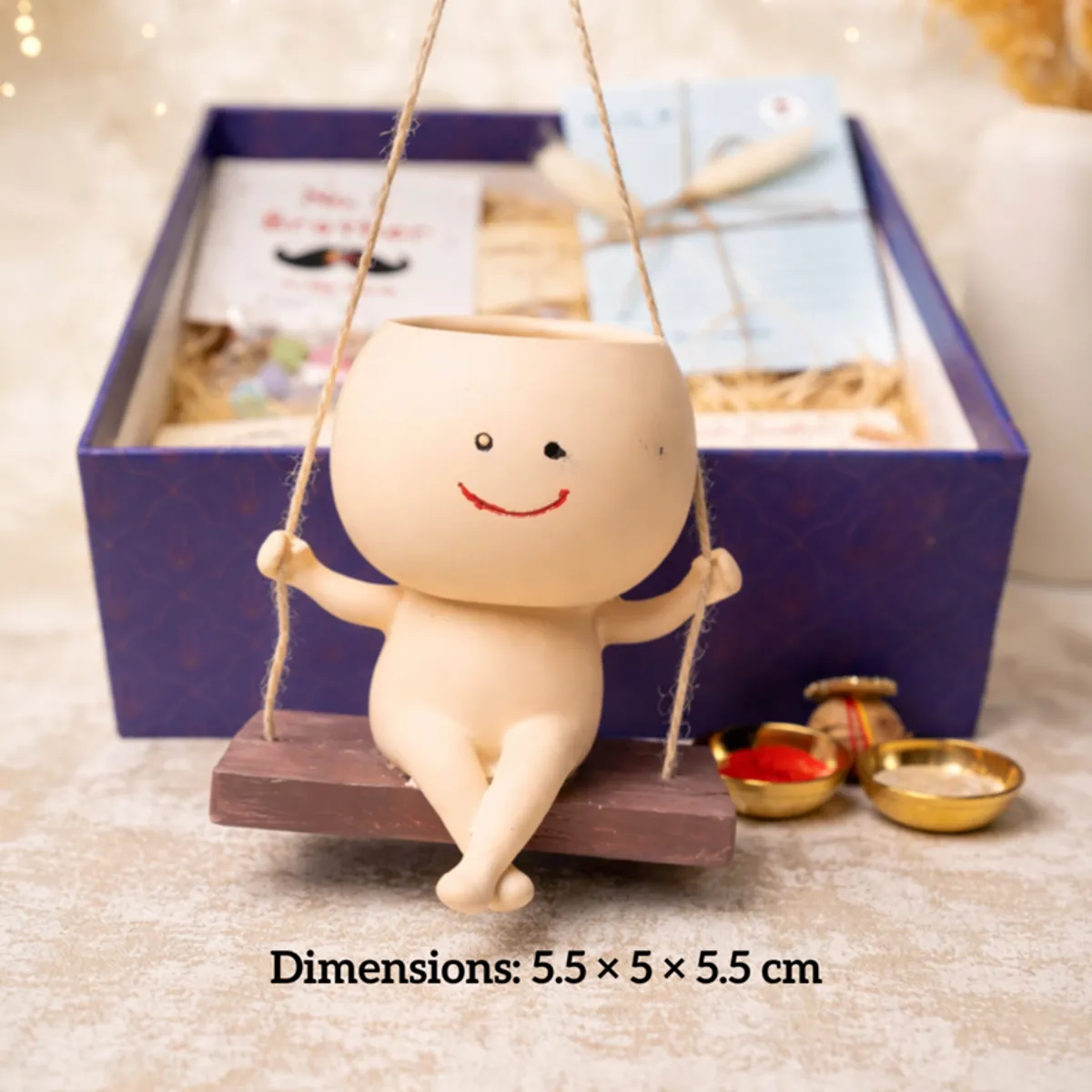 Smiling character planter on a swing beside the open blue Rakhi hamper