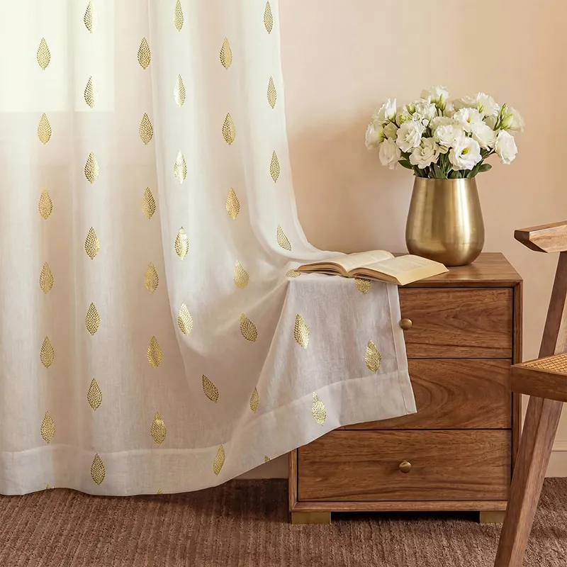 White curtain with gold leaf motifs draped beside a wooden side table and vase
