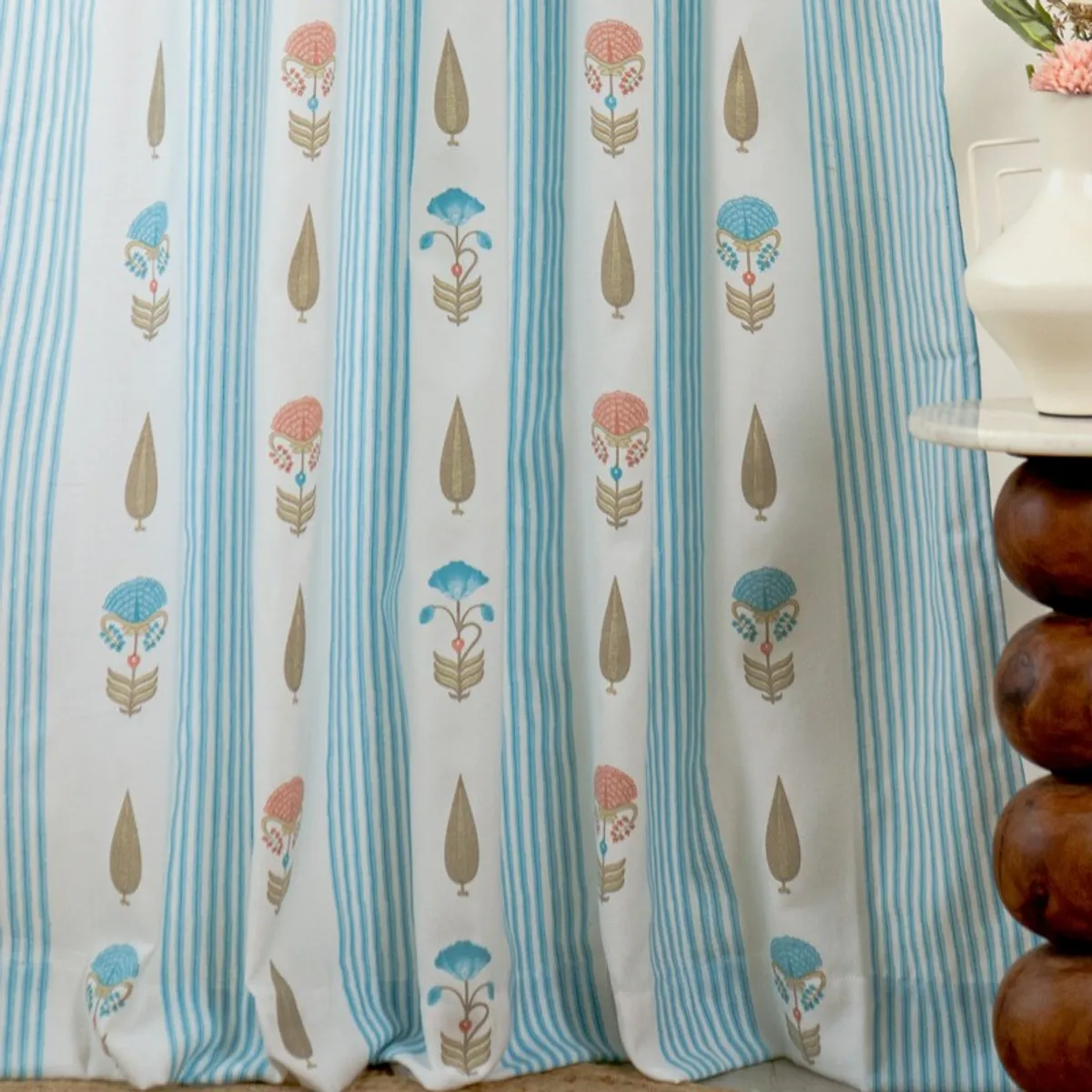 Blue and white patterned curtain hanging beside a white vase and wooden table