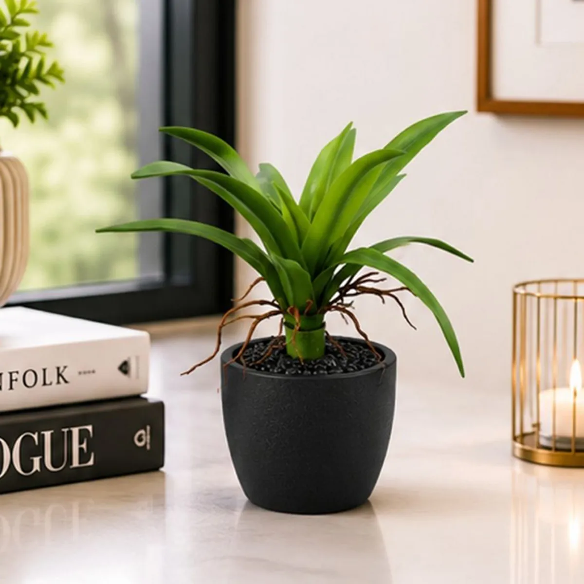Green artificial bromeliad with long leaves shown on a tabletop beside books and a candle