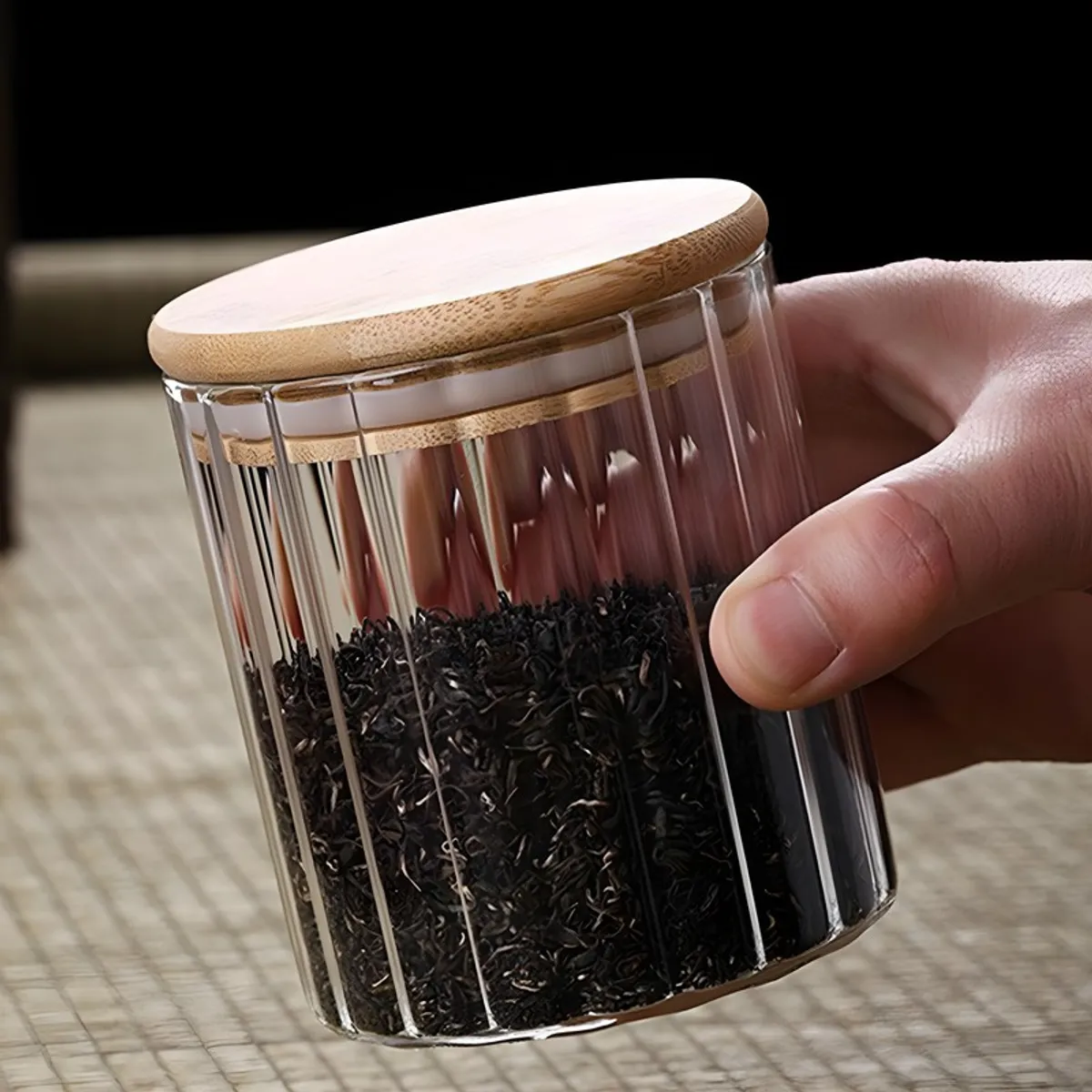 Hand holding a clear fluted storage jar filled with tea leaves.
