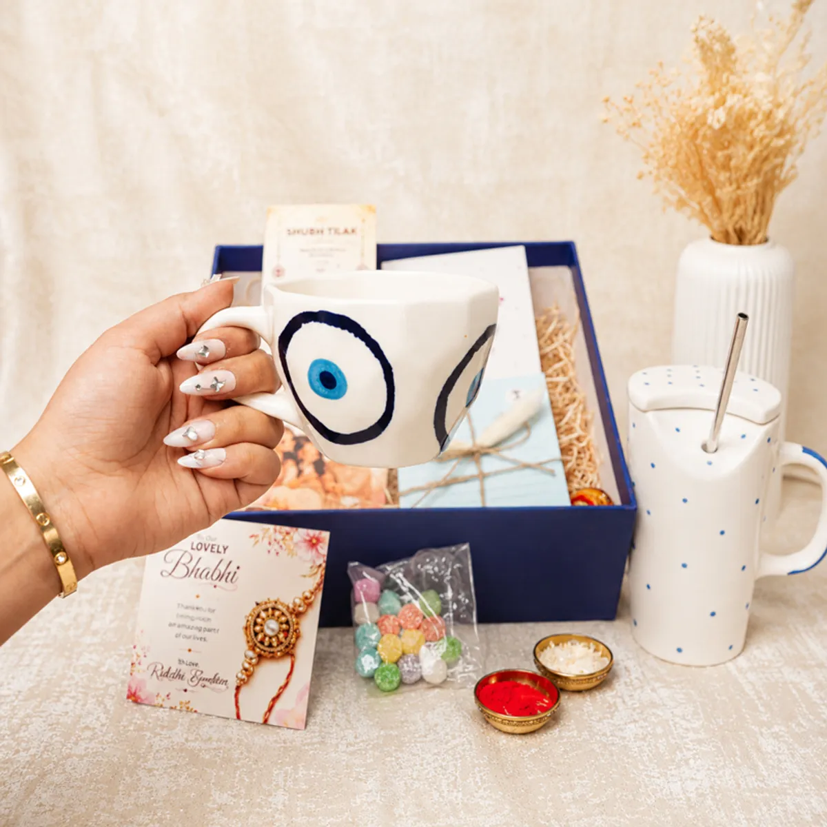 Hand holding the evil eye mug beside the blue tumbler and hamper cards.