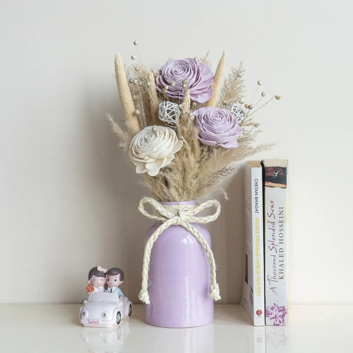 Dried flower bunch in a lavender vase displayed beside books and a figurine.