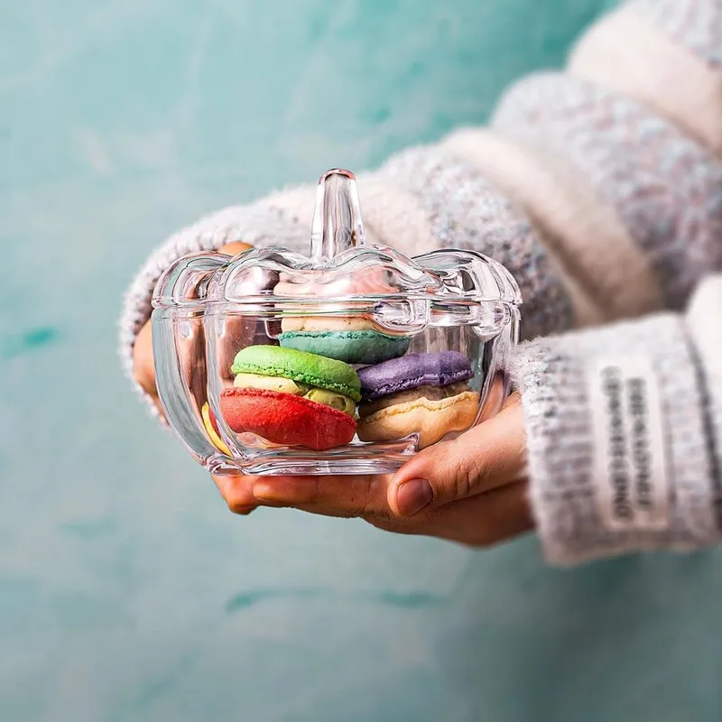 Transparent lidded glass bowl filled with colourful macarons held in both hands