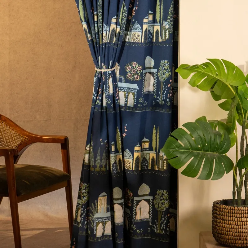 Blue printed curtain tied back beside a wooden chair and leafy plant.