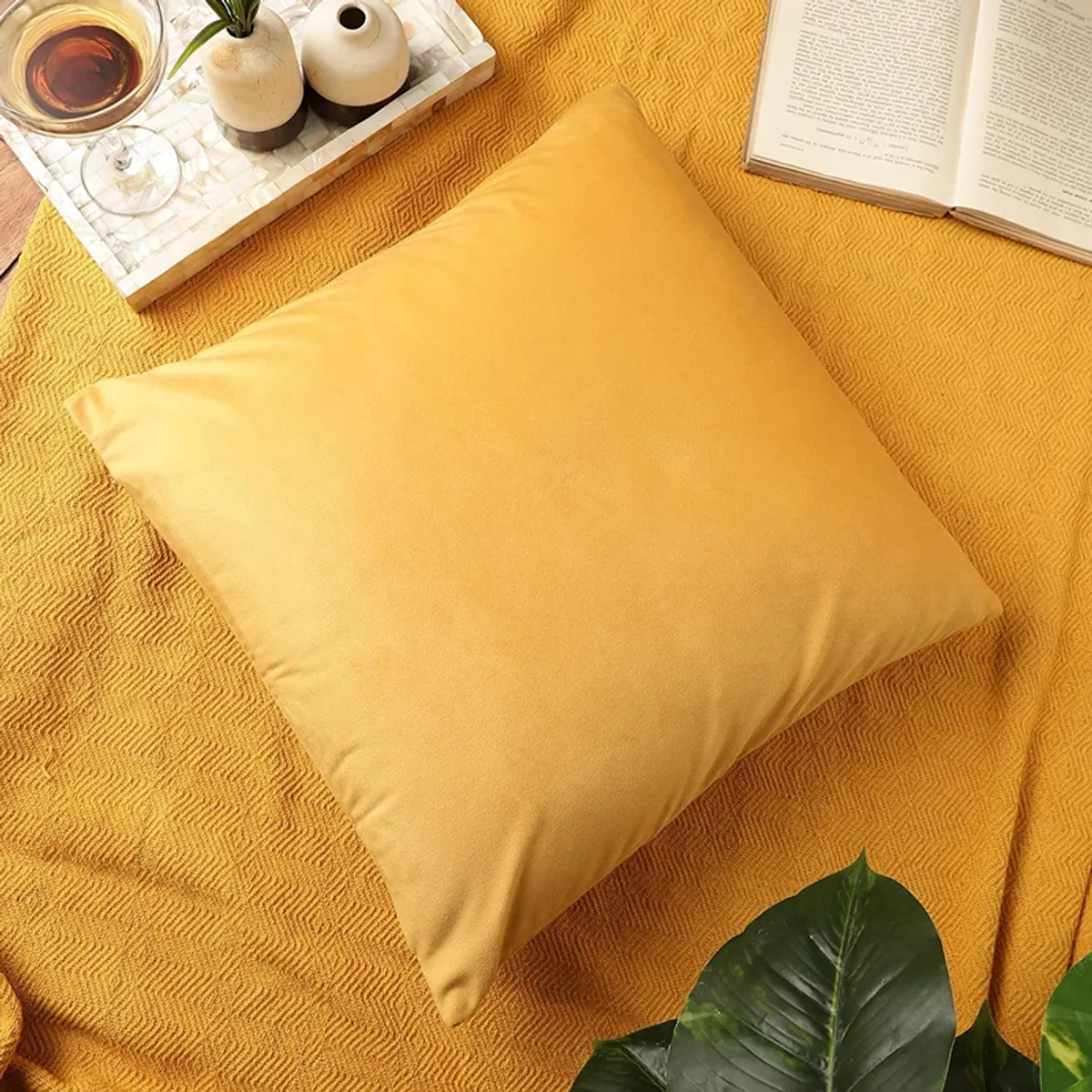Yellow square cushion cover styled on a textured surface with a book and tea