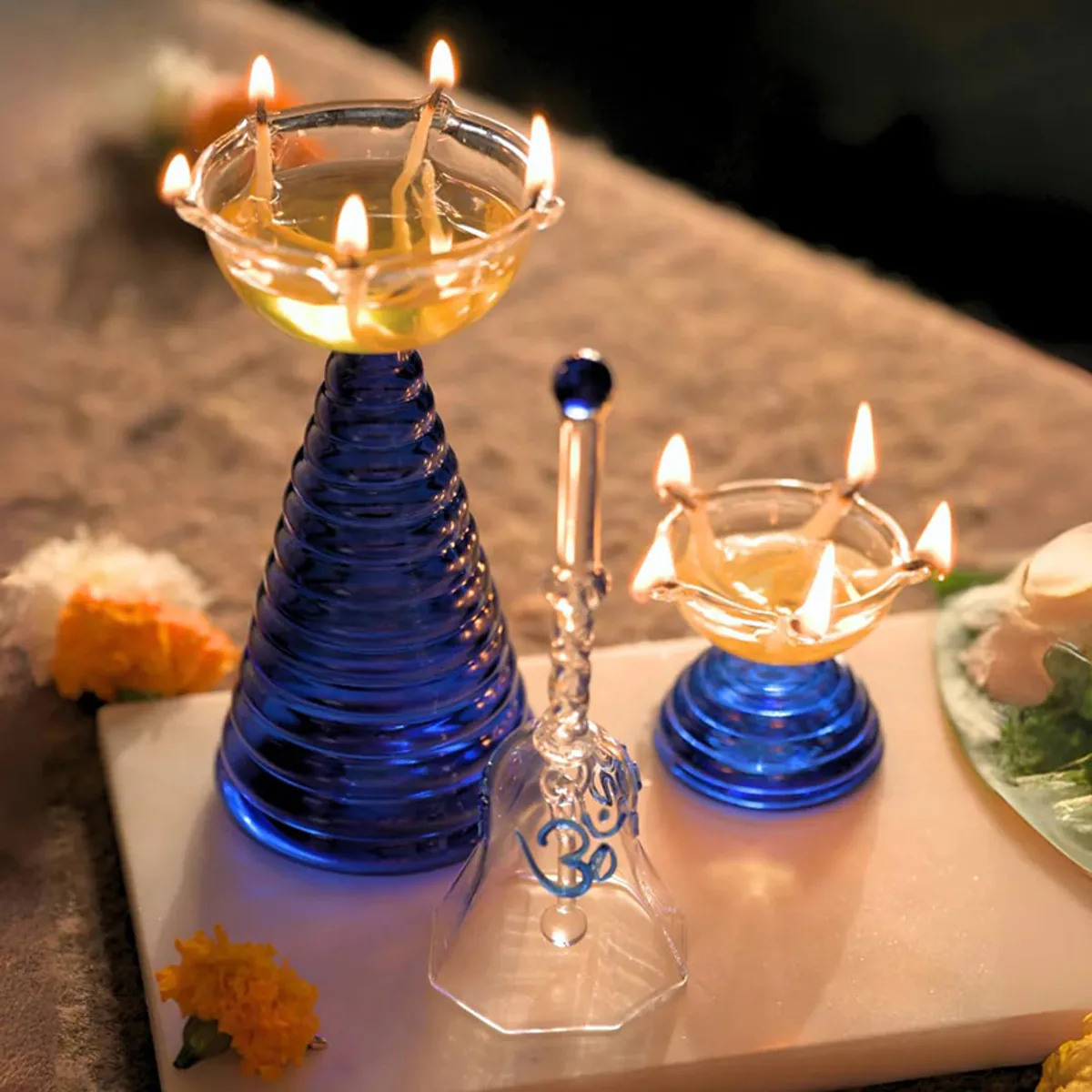 Blue glass diya pair and transparent pooja bell displayed with lit diyas