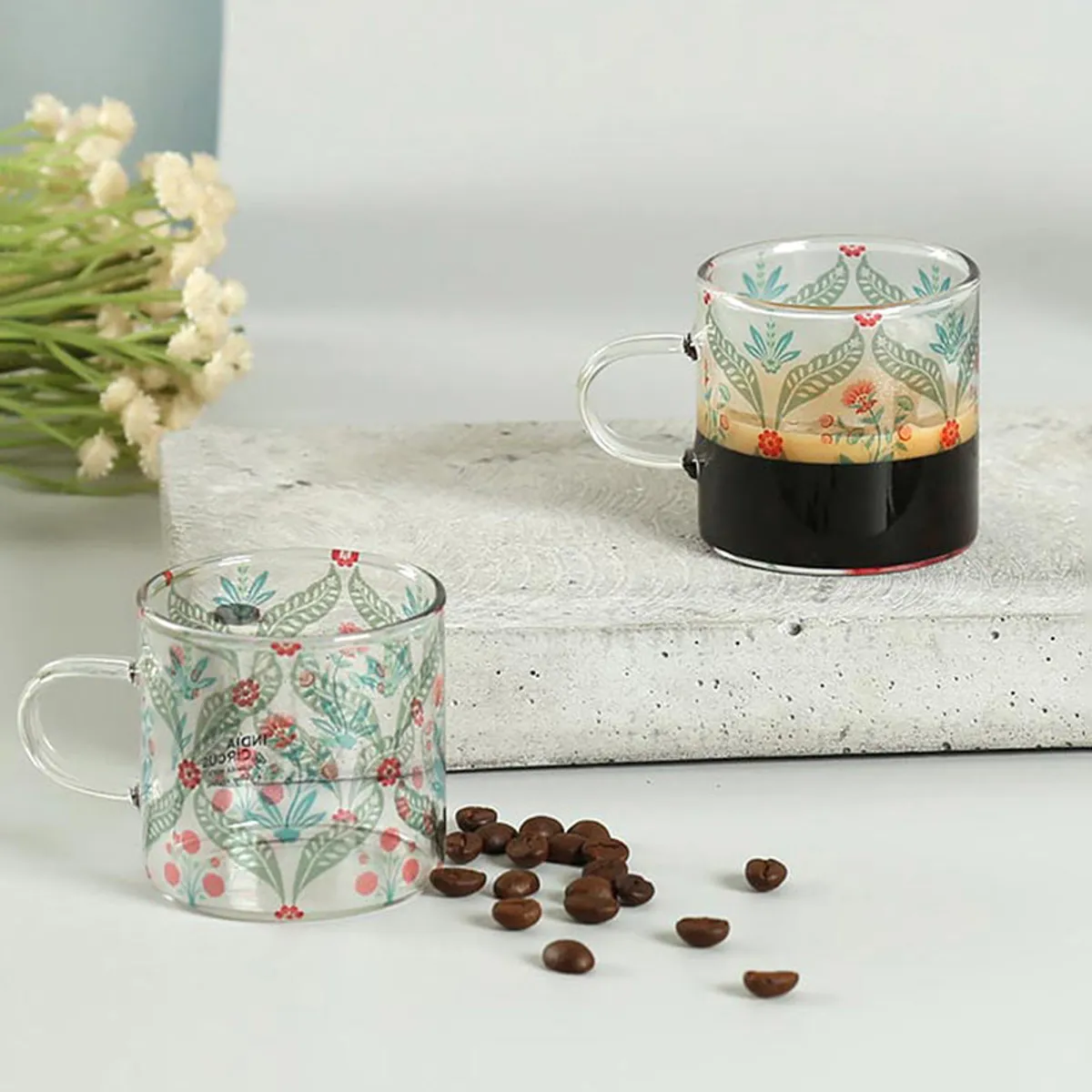 Two floral glass mugs styled with coffee beans on a stone surface