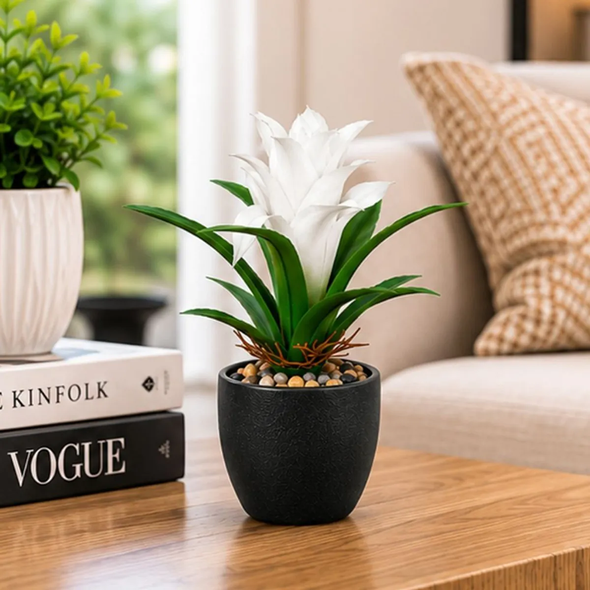 White artificial bromeliad with green leaves in a black planter on a wooden table