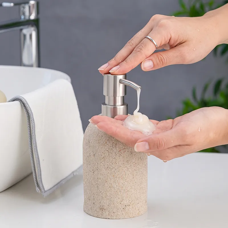 Hand pressing a beige soap dispenser with silver pump beside a sink