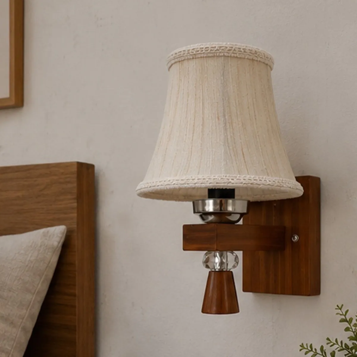 White pleated wall lamp with brown wooden base shown beside a bed