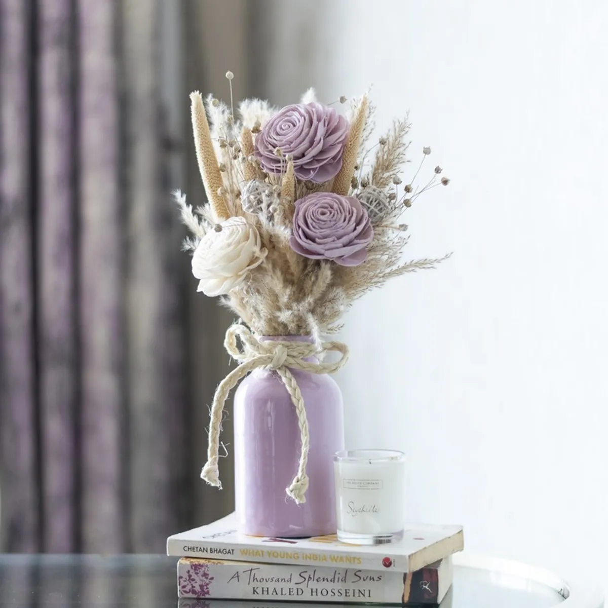 Dried flower bunch arranged in a lavender vase beside a candle and books.