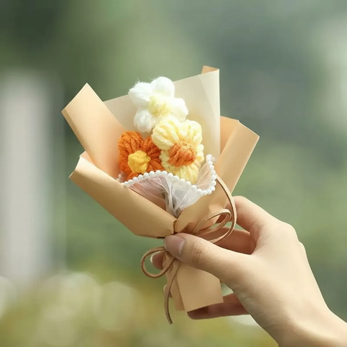 Hand holding a small beige-wrapped crochet flower arrangement with white, yellow and orange flowers.
