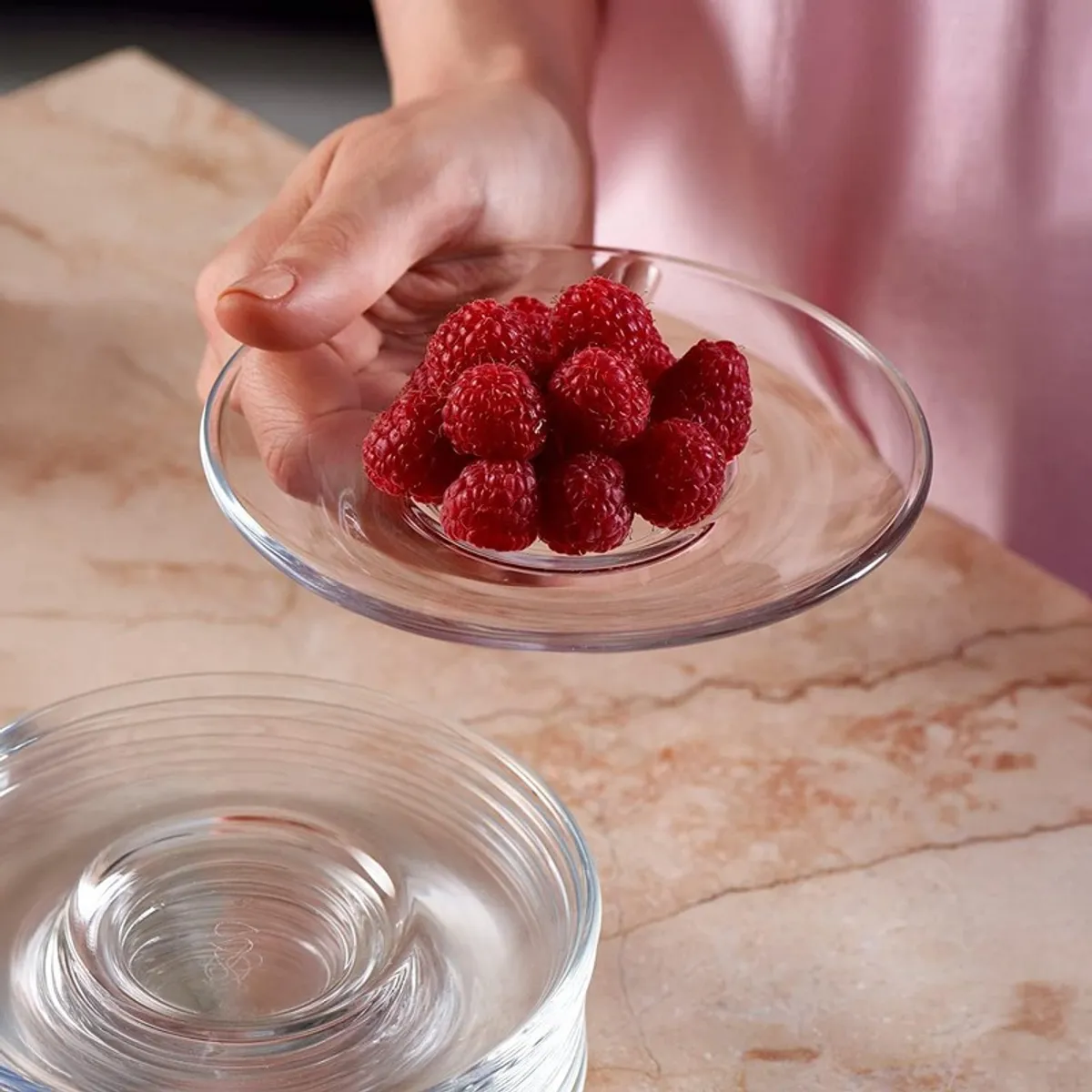 Hand holding a clear glass saucer filled with raspberries