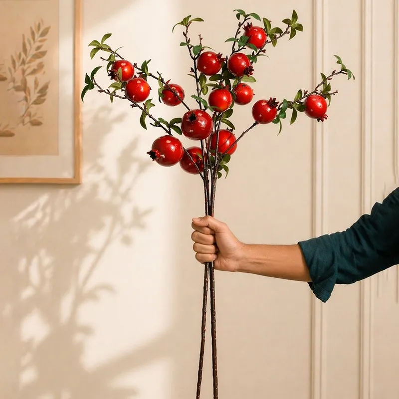 Hand holding tall artificial pomegranate branches with red fruit and green leaves