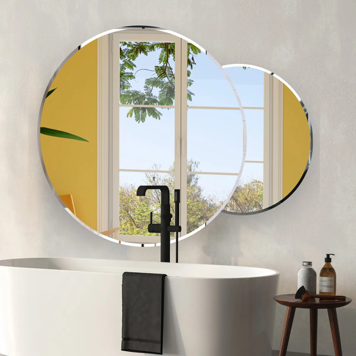 Round silver wall mirror above a bathtub reflecting a bright window and greenery