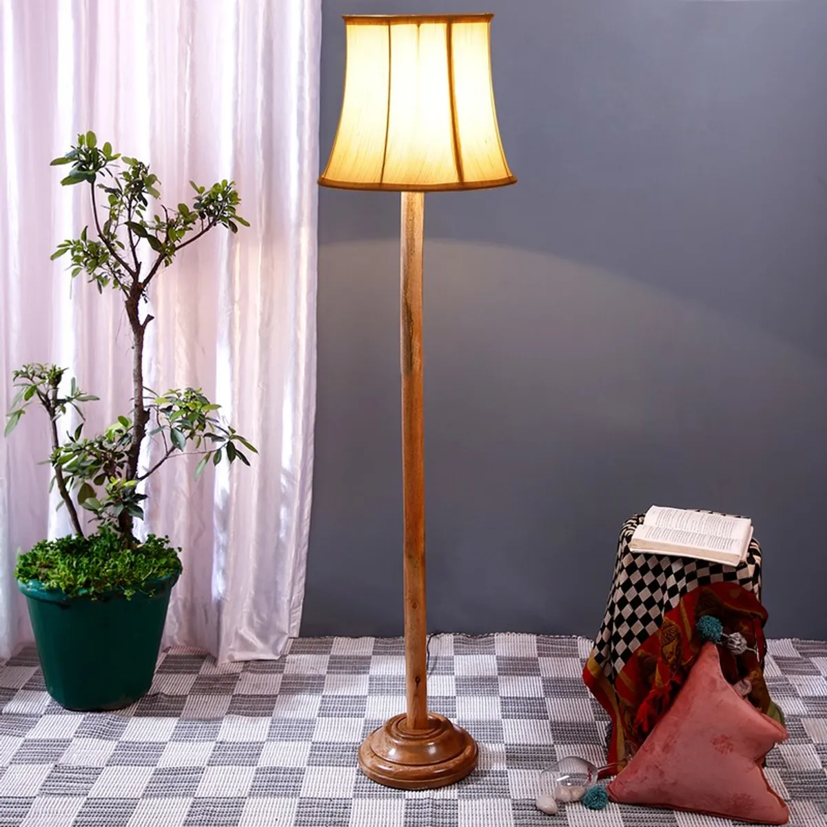 Wooden floor lamp with lit white shade placed beside a potted plant and draped stool