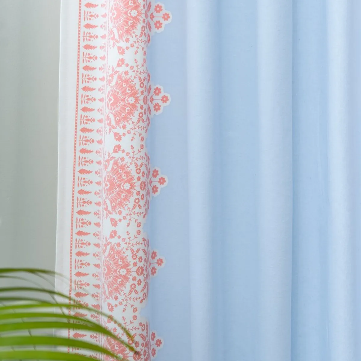 Blue curtain panel with pink floral edging beside a leafy plant