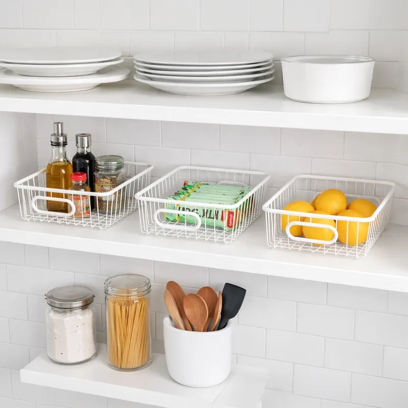 Three white metal baskets arranged on kitchen shelves with pantry items and fruit