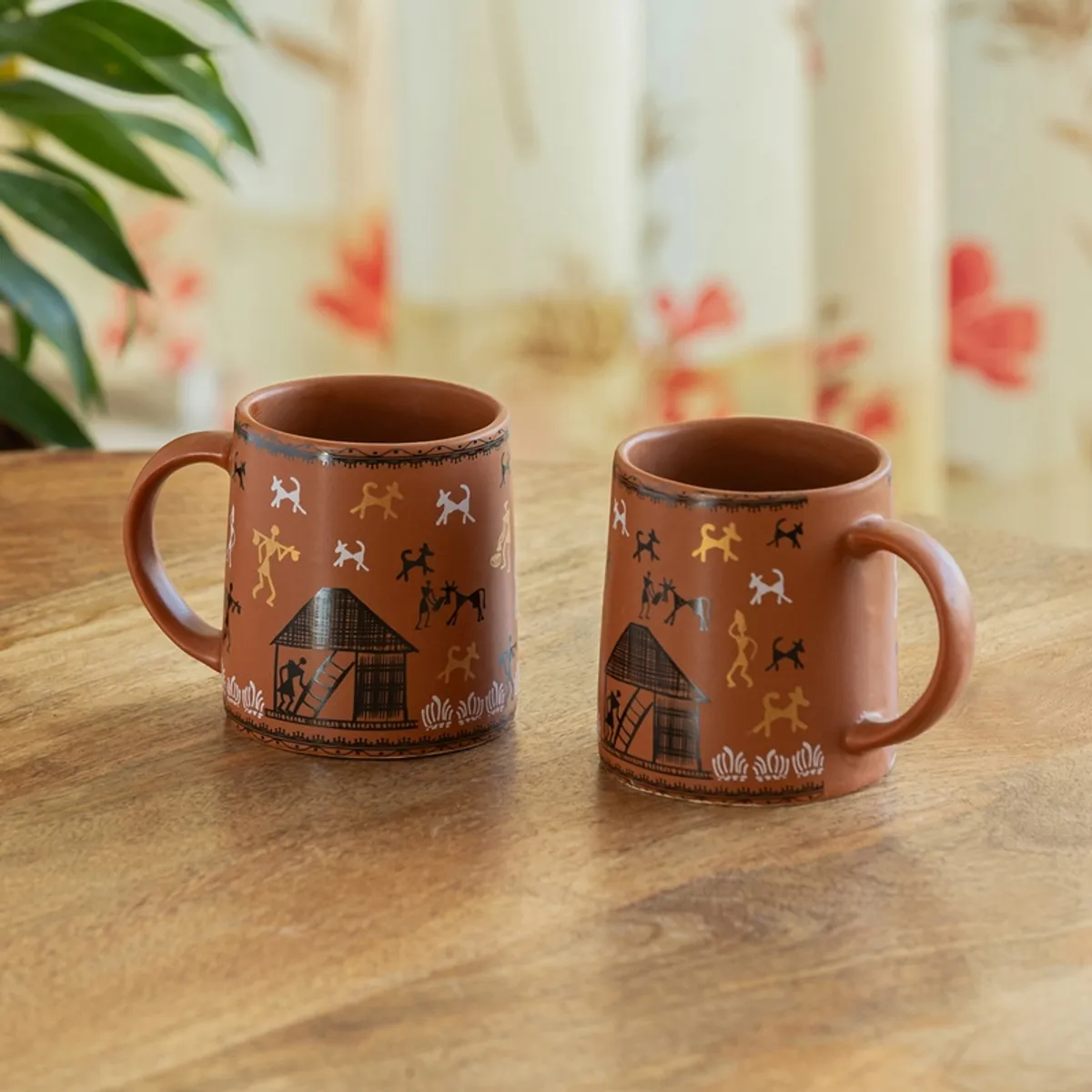 Pair of brown ceramic mugs with colourful Warli figures on a wooden table.