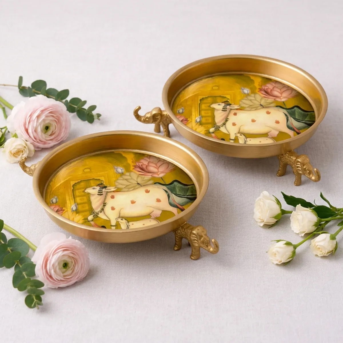 Two round gold Urlis with elephant bases and colourful floral artwork on a white backdrop