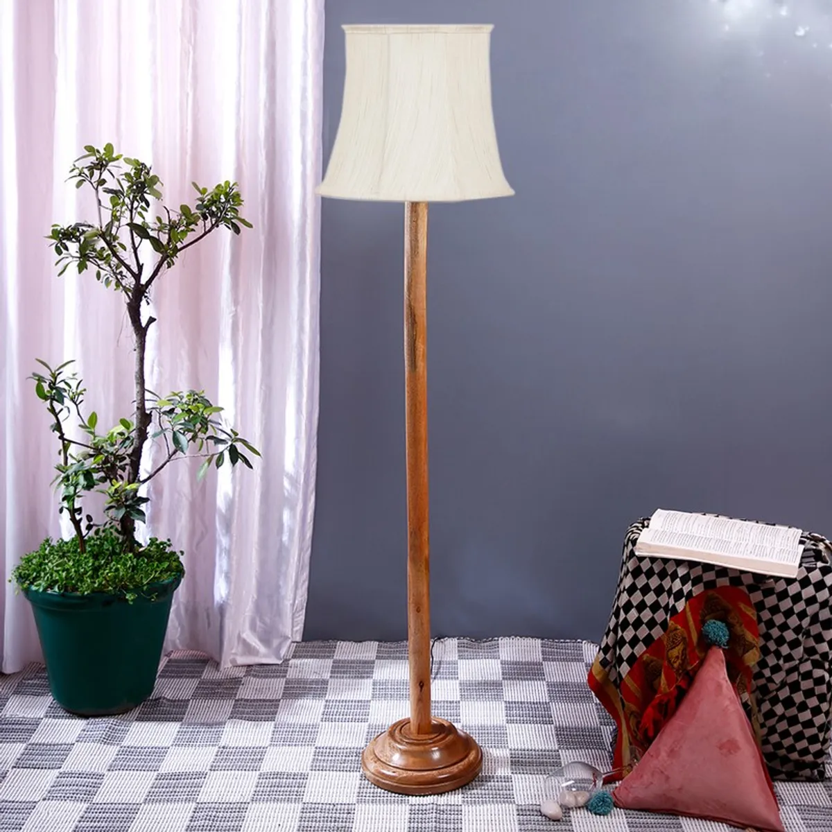 Wooden floor lamp with unlit white shade beside a potted plant and patterned stool