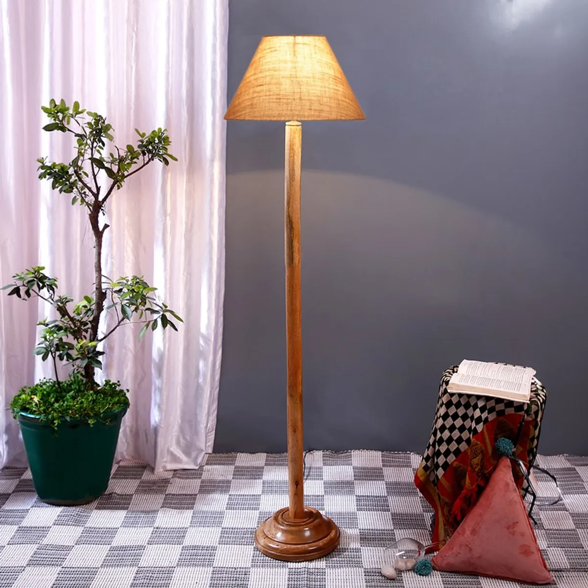 Wooden floor lamp with lit jute shade styled near a potted plant and draped stool