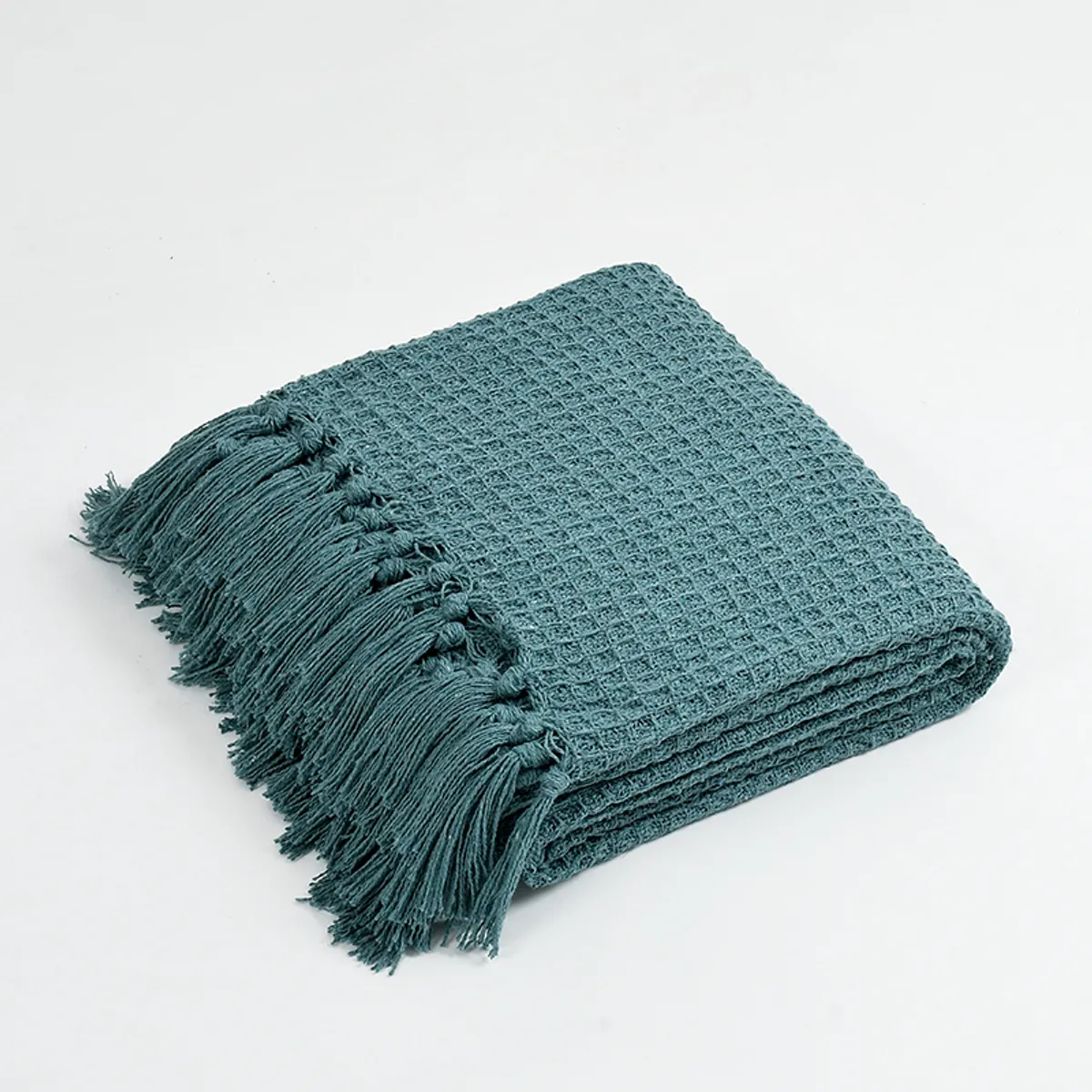 Folded teal waffle throw with tasselled fringe on a white background