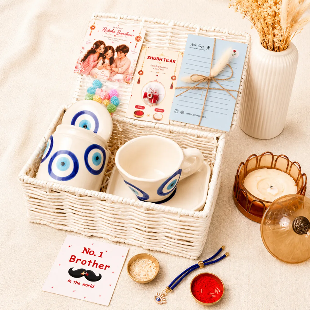 Open white basket containing evil eye drinkware, Raksha Bandhan cards, sweets and ritual items