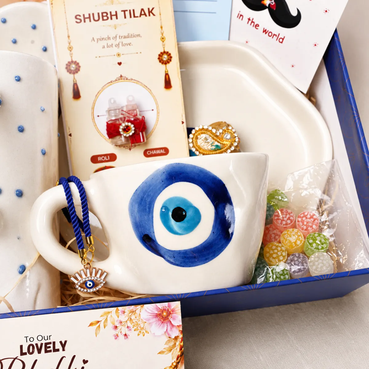 Close-up of evil eye mug, rakhi, festive card and colourful sweets in the hamper.